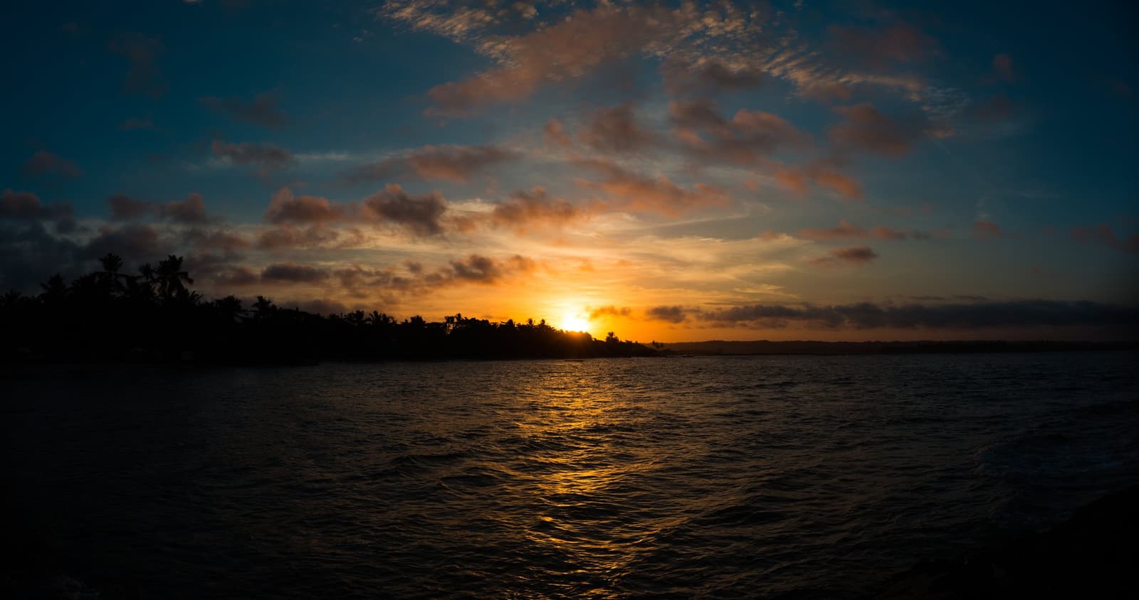 The sea goes quiet, and the sky does the talking. Golden sunset reflecting across calm ocean water, with a dark silhouette of palm-lined coast on the horizon, seen from the water during an itacaré surf school session
