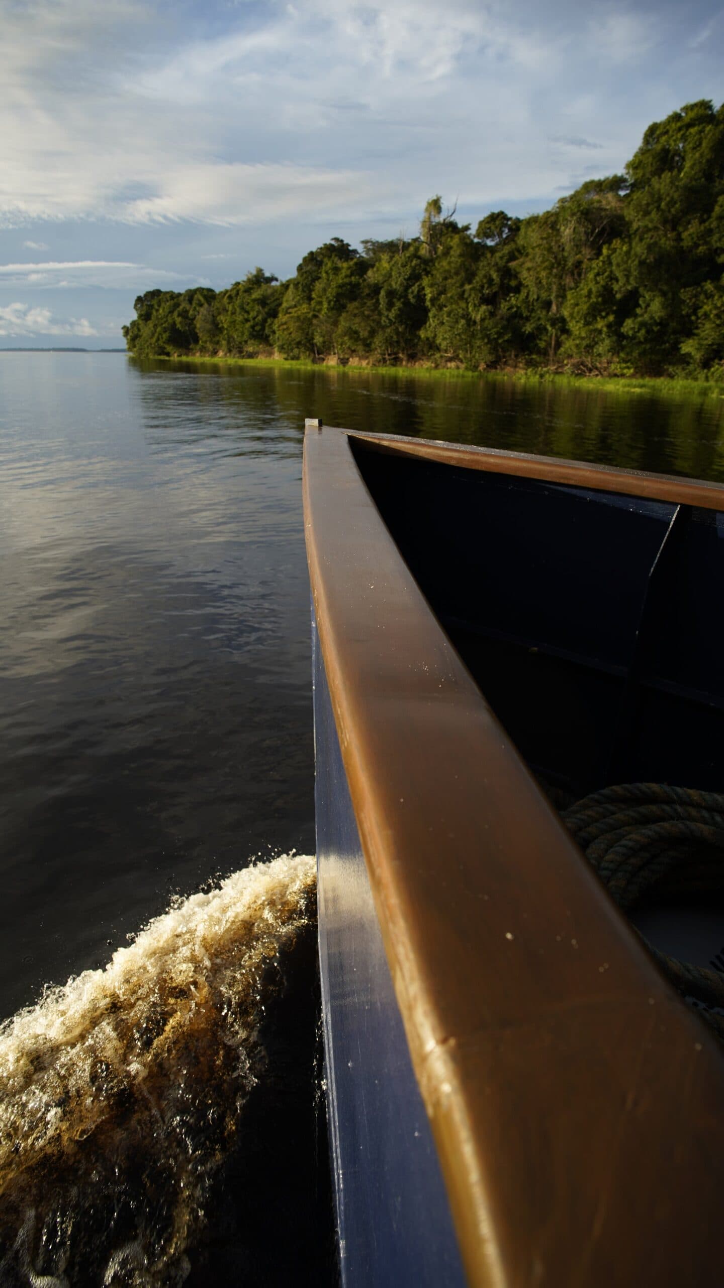 On the water, the forest takes over, and the city feels very far away. View from the bow of a wooden-hulled boat on the la jangada amazon cruise, with dark-water river and dense rainforest lining the far bank under a partly cloudy sky