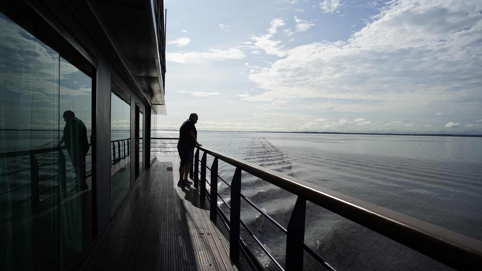 The wake behind you is the only proof the river was ever still. A silhouetted guest leans on the railing of La Jangada amazon cruise's open deck, watching the wide river wake trail into the distance under a partly cloudy sky
