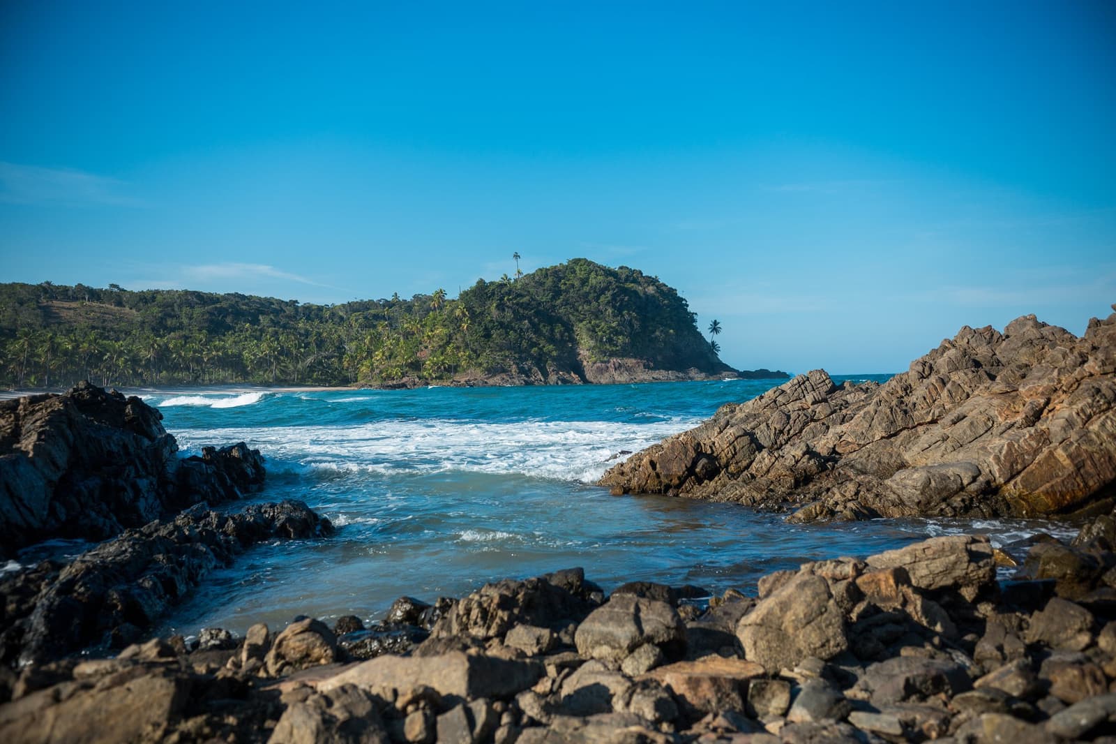Rocky points, lush hills, warm water, this is the classroom. Jagged dark rocks frame a cove at the itacaré surf school coastline, with turquoise waves rolling toward a sandy beach beneath a forest-covered headland and clear blue sky