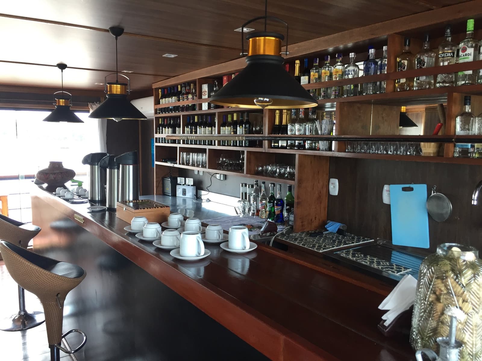 Coffee before the dolphins, sundowners after. The bar has you covered either way. Polished wood bar counter on the La Jangada amazon cruise with white espresso cups, a thermal coffee dispenser, and shelves stocked with spirits, wines, and glassware beneath black pendant lights