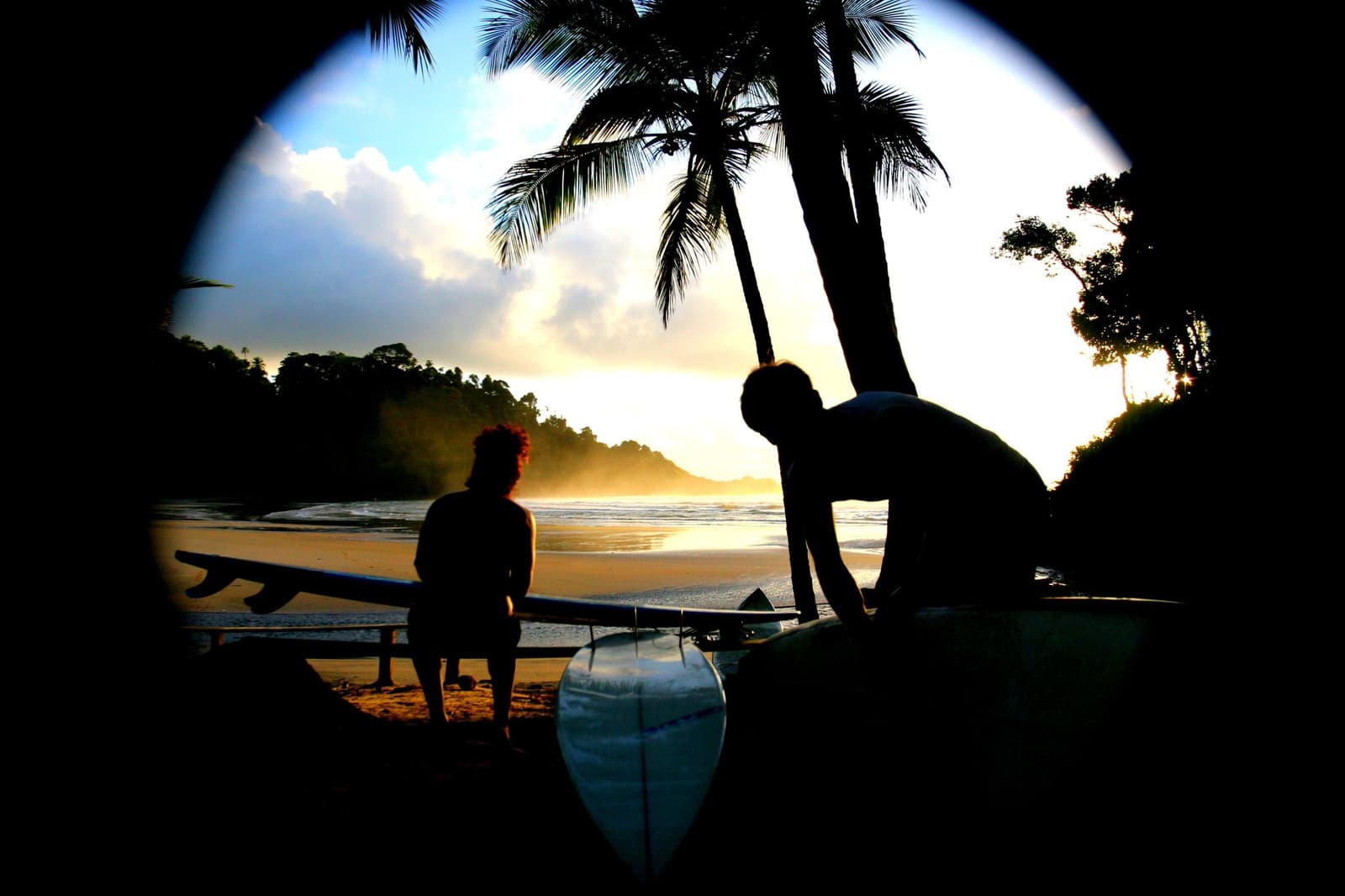 Last light, boards ready, and the ocean still has a few waves to give. Photo: Picasa Two surfers silhouetted against a golden sunset on a tropical beach in Itacaré surf school, waxing and checking longboards beside palm trees with forested headlands in the background