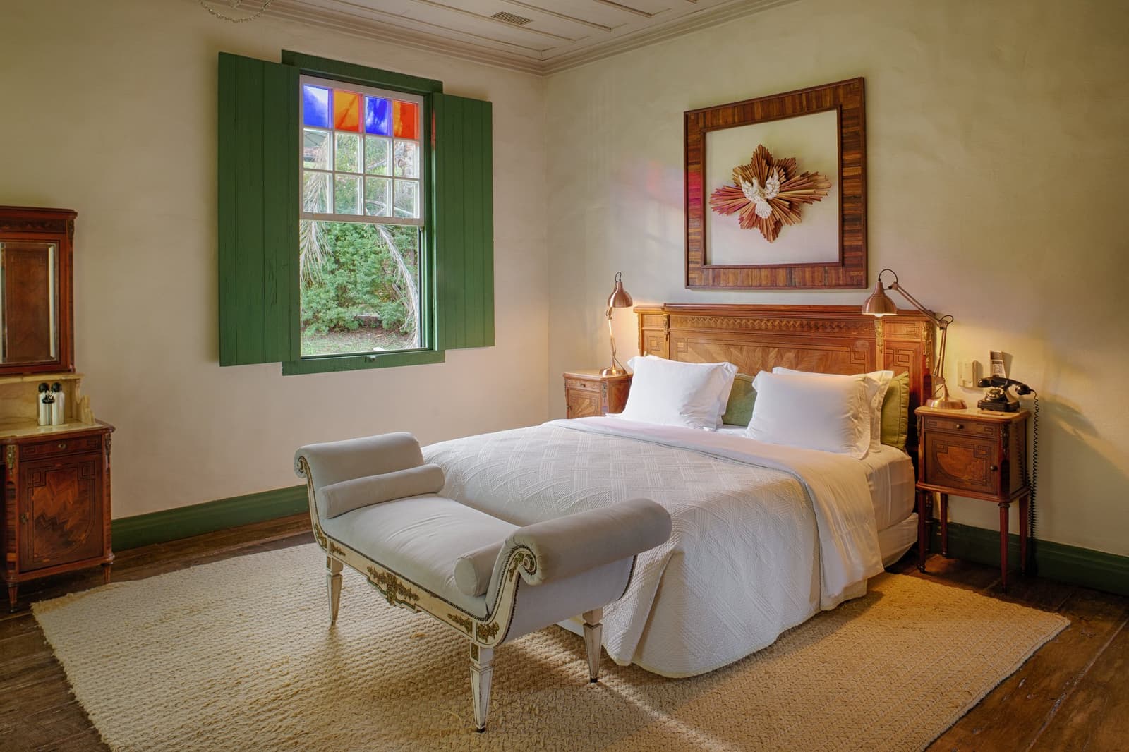 Old farmhouse bones, fresh white linen. Sleep well, you have a canyon in the morning. Photo: Henrique Mangeon A warmly lit bedroom at the Ibiti Project's Engenho Lodge, featuring a carved wooden headboard, crisp white bedding, an ornate upholstered bench, green shuttered window with stained-glass panels, and antique timber furniture on wide-plank floors