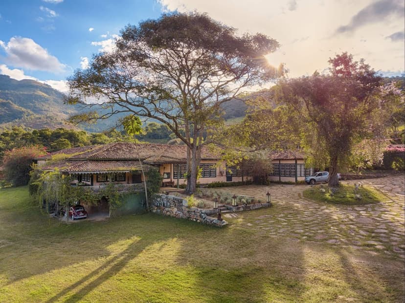 Aerial view of the Ibiti Project's Engenho Lodge, a colonial-style farmhouse with terracotta roof tiles, a wide veranda, and a cobblestone courtyard, framed by a large spreading tree and green mountain ridges in the background Aerial view of the Ibiti Project's Engenho Lodge, a colonial-style farmhouse with terracotta roof tiles, a wide veranda, and a cobblestone courtyard, framed by a large spreading tree and green mountain ridges in the background