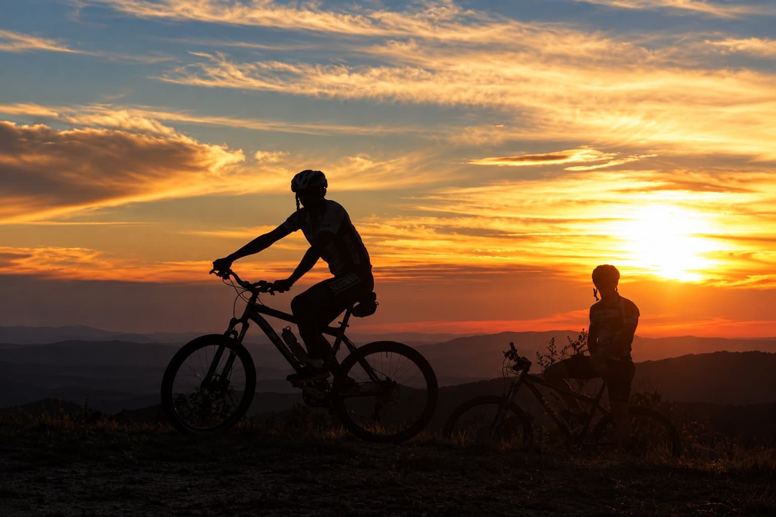 Good legs help, but the view does most of the work. Two cyclists silhouetted against a fiery orange and red sunset over layered mountain ridges on an Ibiti Project trail ride