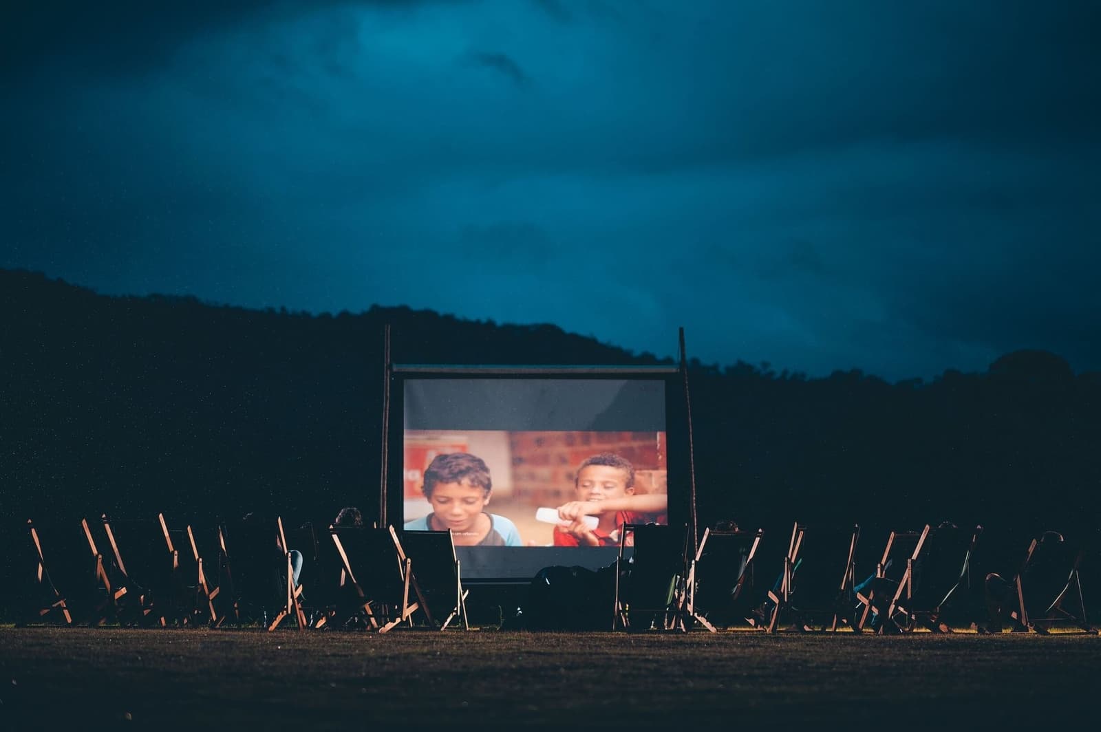 Movies under the clouds, forest at your back, no Wi-Fi needed. Rows of empty wooden folding chairs face an outdoor cinema screen glowing in the dark, showing two children in a film scene, with a forested hillside and deep blue cloudy sky behind, part of the Ibiti Project experience in Minas Gerais.