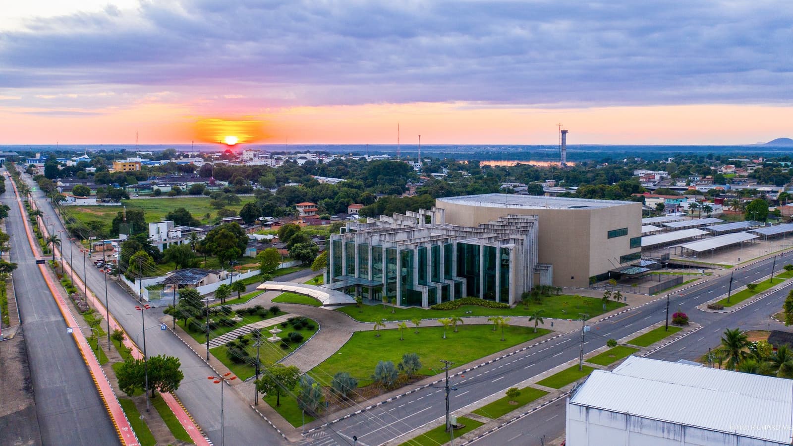 Boa Vista at dusk: the city that sends you off toward one of the oldest mountains on Earth. Aerial view of Boa Vista city at sunset, with a modern glass-fronted civic building surrounded by green lawns, wide avenues, and the city spreading toward the horizon under a pink and orange sky — the starting point of a mount roraima expedition.