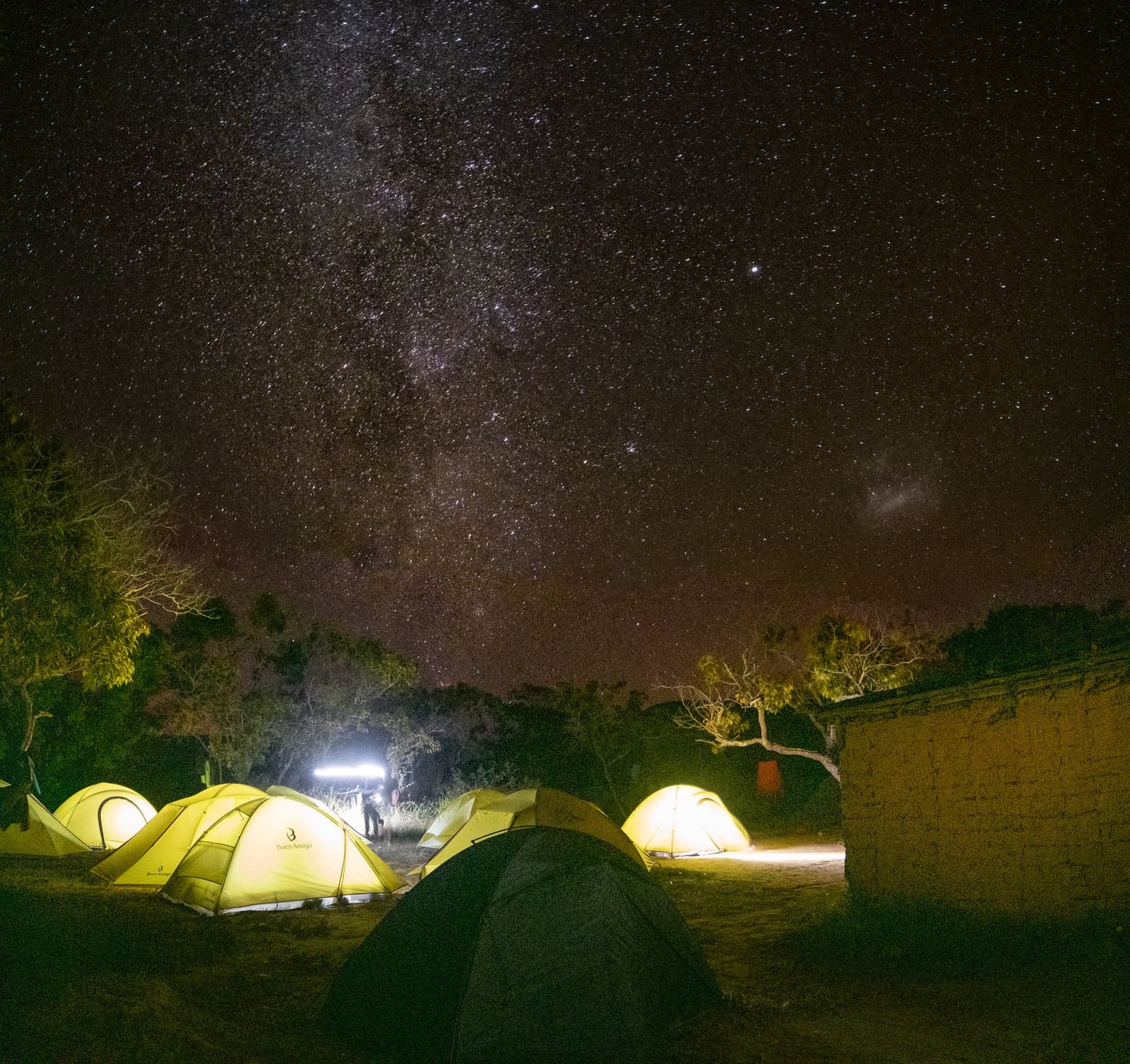 No light pollution, no bad nights. Just the galaxy doing its thing. Glowing yellow dome tents pitched at a wilderness campsite on a mount roraima expedition, with the Milky Way arching across a star-filled sky above sparse trees and a mud wall