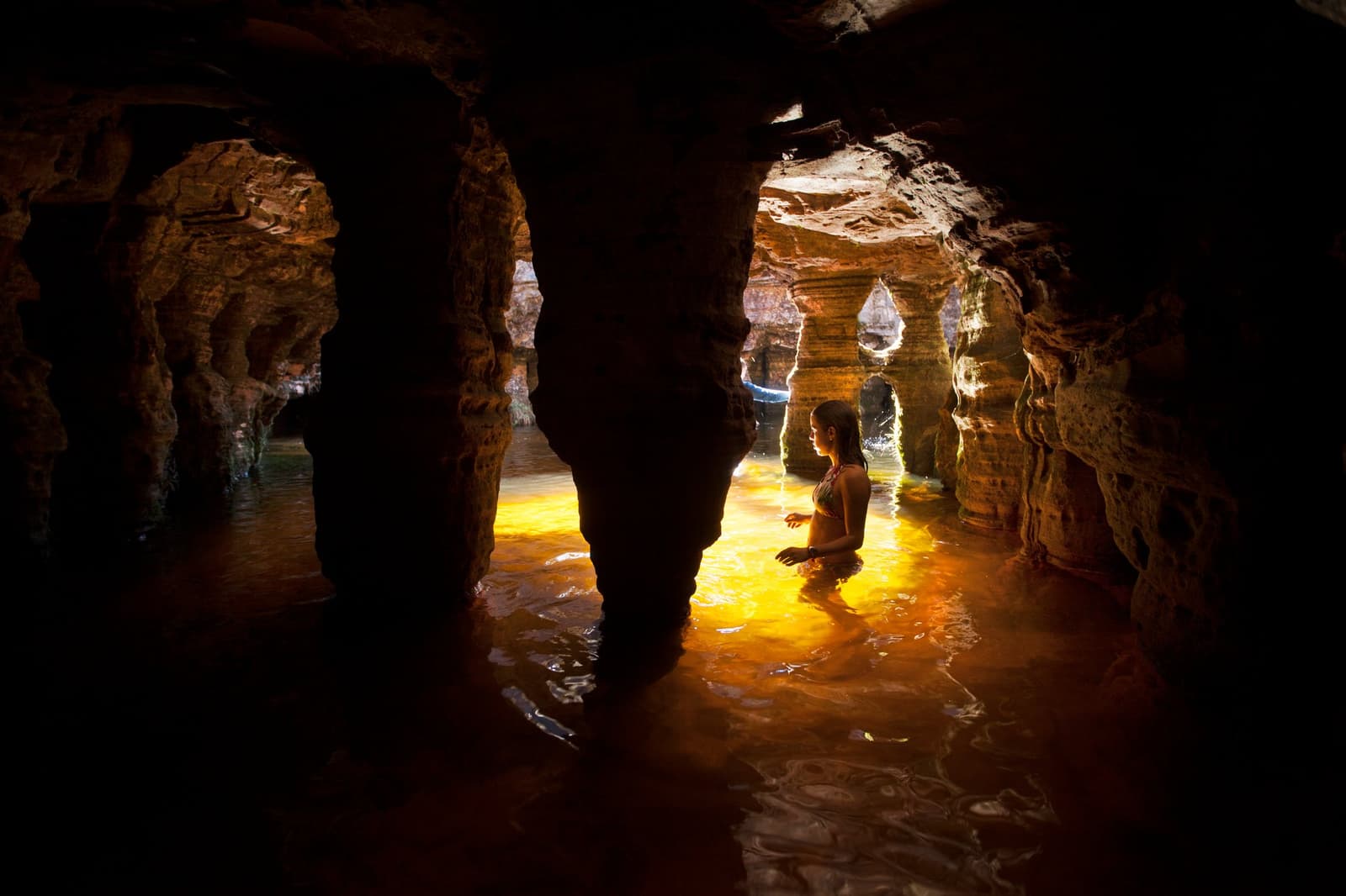 Cool water, warm light, and a cave that took two billion years to carve. A woman wading waist-deep through a flooded sandstone cave on a mount roraima expedition, golden light reflecting off the amber-colored water between sculpted rock columns