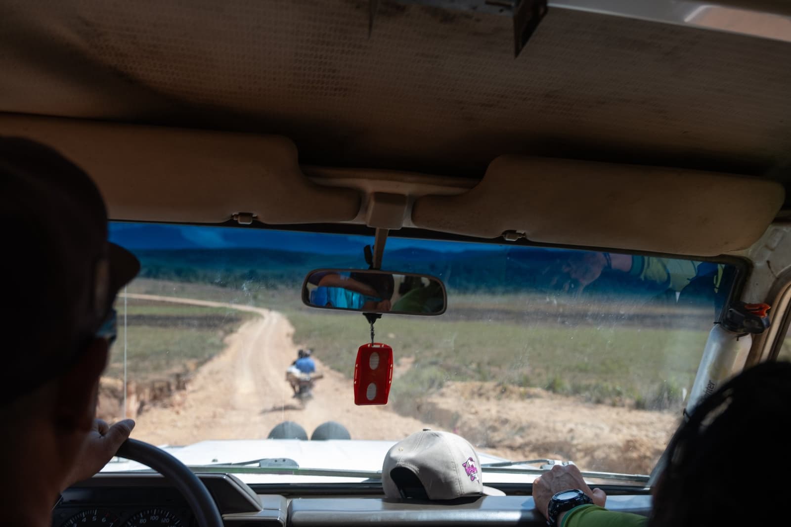 Dirt roads, a dangling key fob, and somewhere ahead, the mountain. View from inside a 4x4 vehicle during a mount roraima expedition, looking through the windshield at a winding dirt track across open grassland, with a motorbike visible ahead and passengers reflected in the rearview mirror
