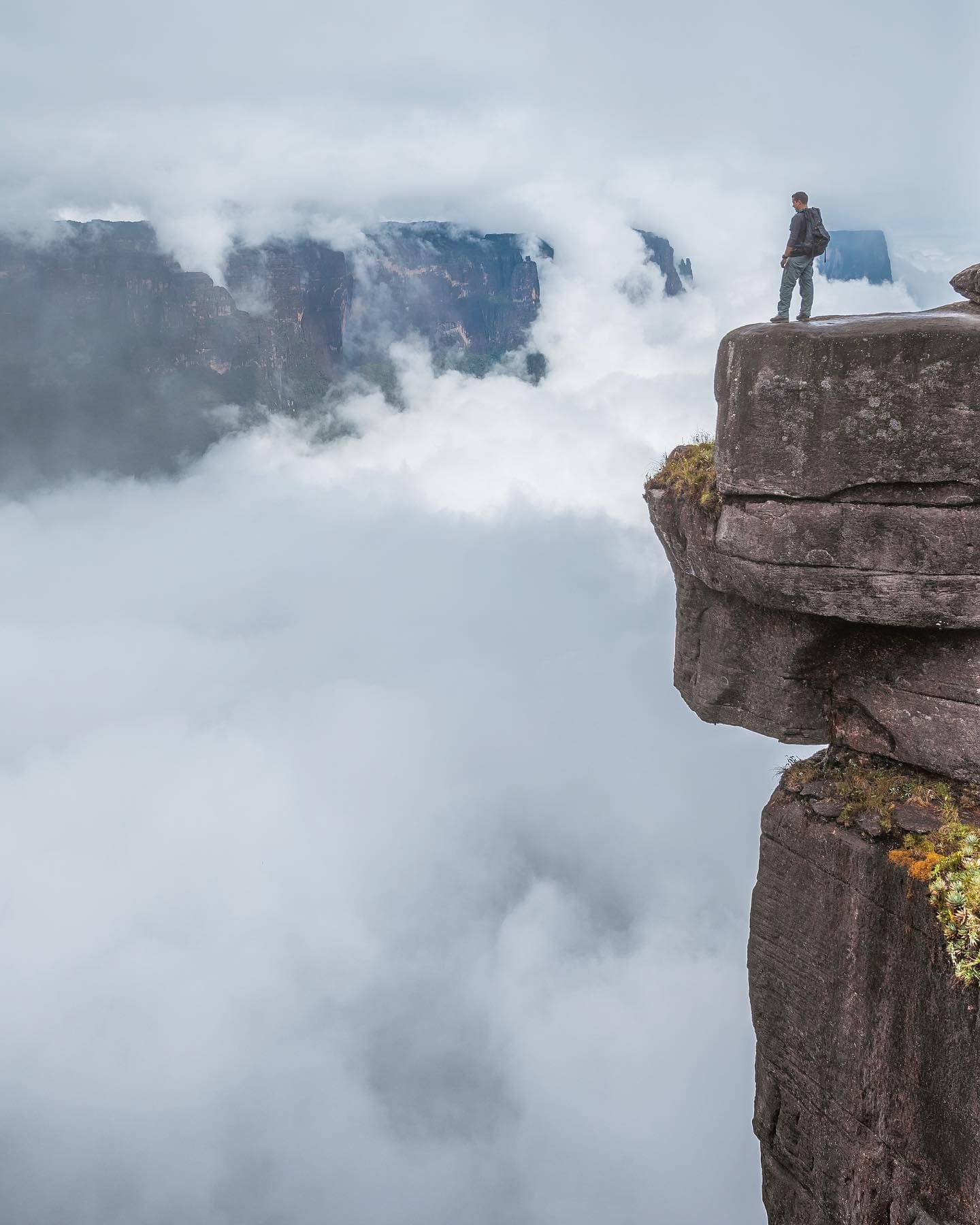 Cloud level is not a figure of speech up here. It is the view. A backpacker stands alone on a narrow ancient rock outcrop during a mount roraima expedition, surrounded by a sea of clouds with a distant tepui wall emerging through the mist