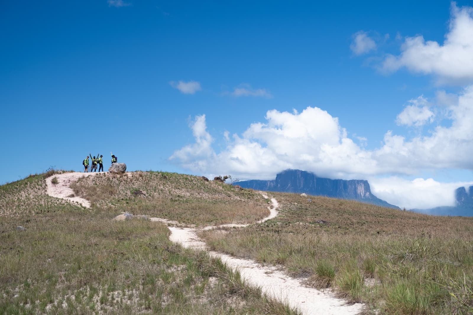 The kind of hill where someone always throws their arms up first. Four hikers celebrating on a sandy hilltop along the mount roraima expedition trail, with the flat-topped tepui visible through clouds in the background