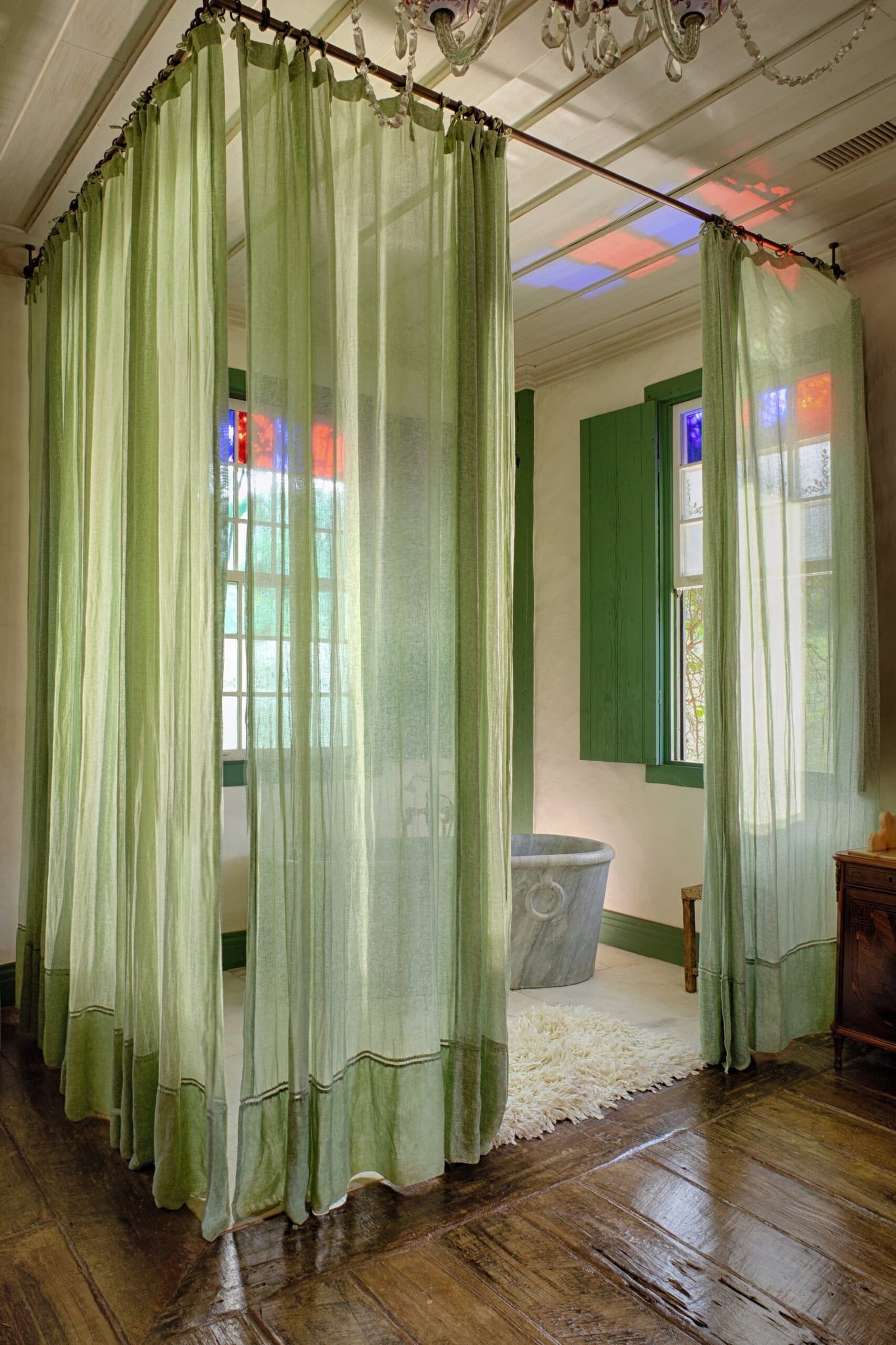 A bathtub framed by stained-glass light and linen curtains. Sleep well tonight. Photo: Henrique Mangeon Sheer sage-green canopy curtains on a four-poster frame in an Ibiti Project lodge room, with a freestanding stone bathtub, green-shuttered windows casting stained-glass light, and polished hardwood floors