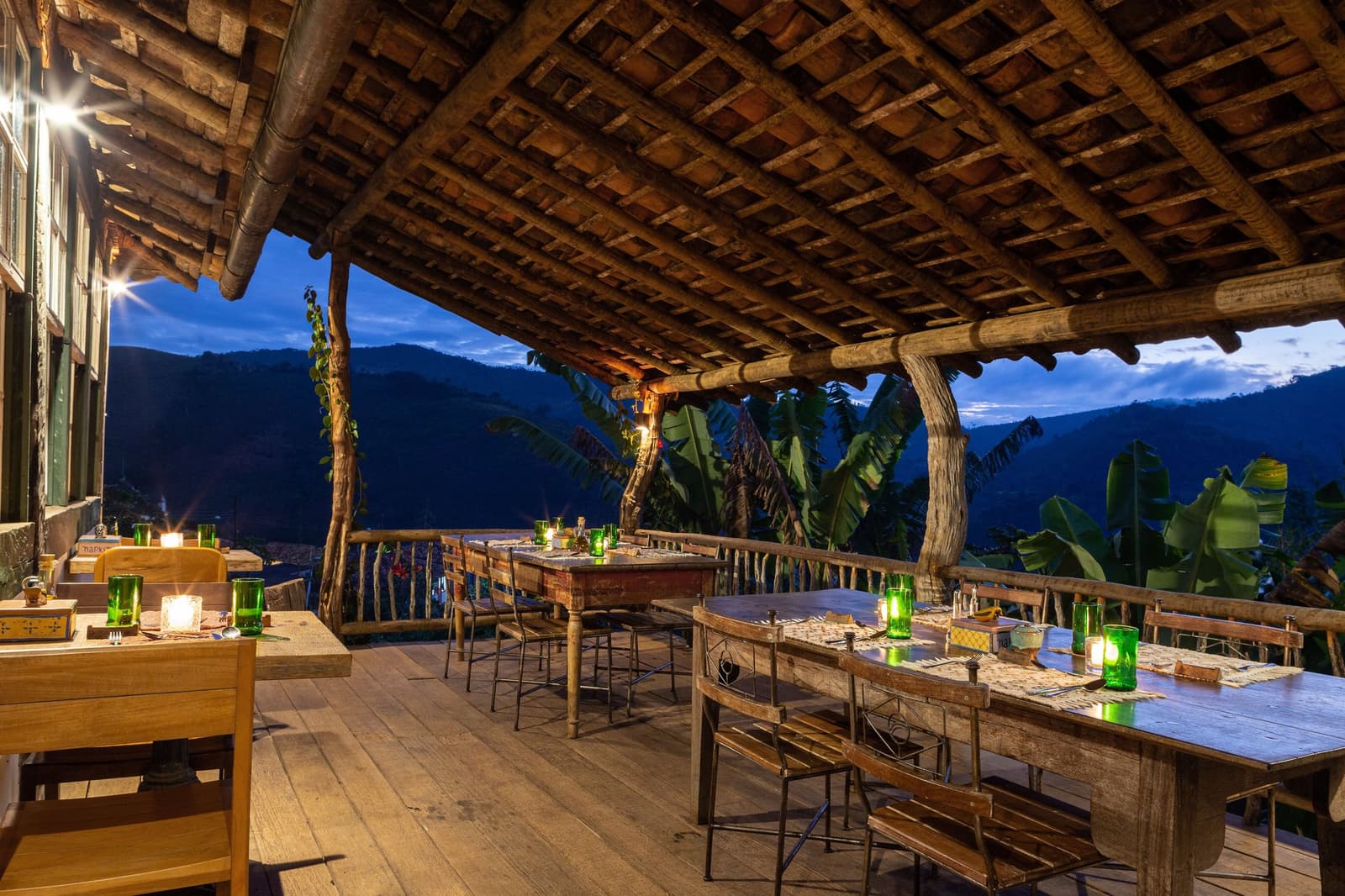 Dinner is served, the valley has dimmed its lights, and the mountains are doing the rest. Candlelit open-air terrace at the Ibiti Project's Engenho Lodge, with rustic wooden tables set for dinner under a timber-beam roof and a twilight mountain valley stretching beyond the railing