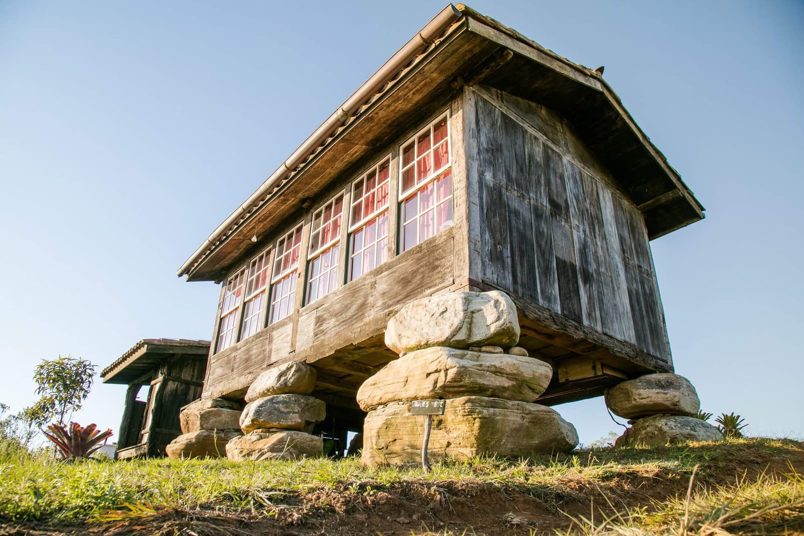 Built to last, long before anyone called it heritage. A weathered wooden building elevated on stacked stone foundations at the Ibiti Project reserve in Minas Gerais, photographed from a low angle against a clear blue sky, with red curtains visible through the multi-pane windows