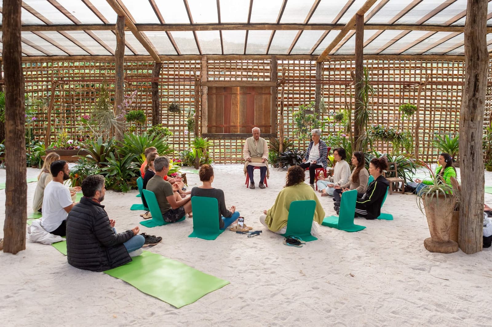 Still minds, sand underfoot, and orchids watching from the walls. A group of travellers sits in a circle on white sand inside a open bamboo-lattice pavilion surrounded by tropical plants, during a guided meditation session at the Ibiti Project in Minas Gerais.