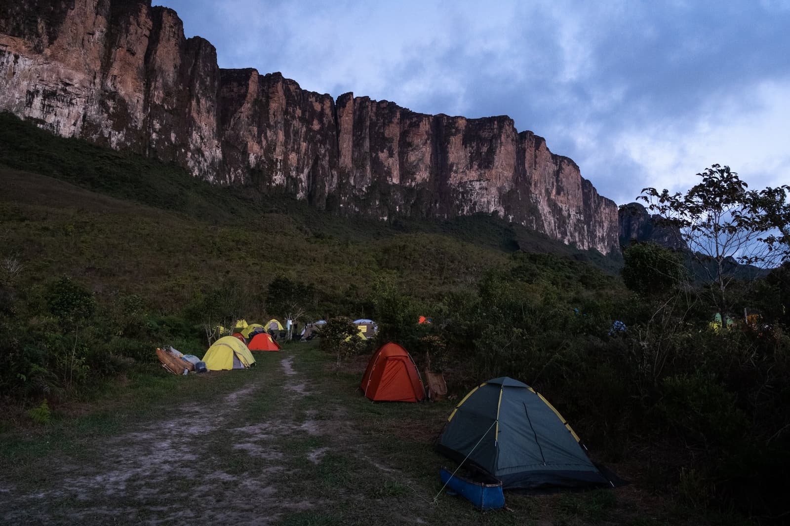 Camp is set, the cliff is close, and tomorrow the real climb begins. Colorful dome tents pitched at dusk on a grassy clearing during a mount roraima expedition, with the towering sandstone cliff wall glowing red under a cloudy blue sky in the background