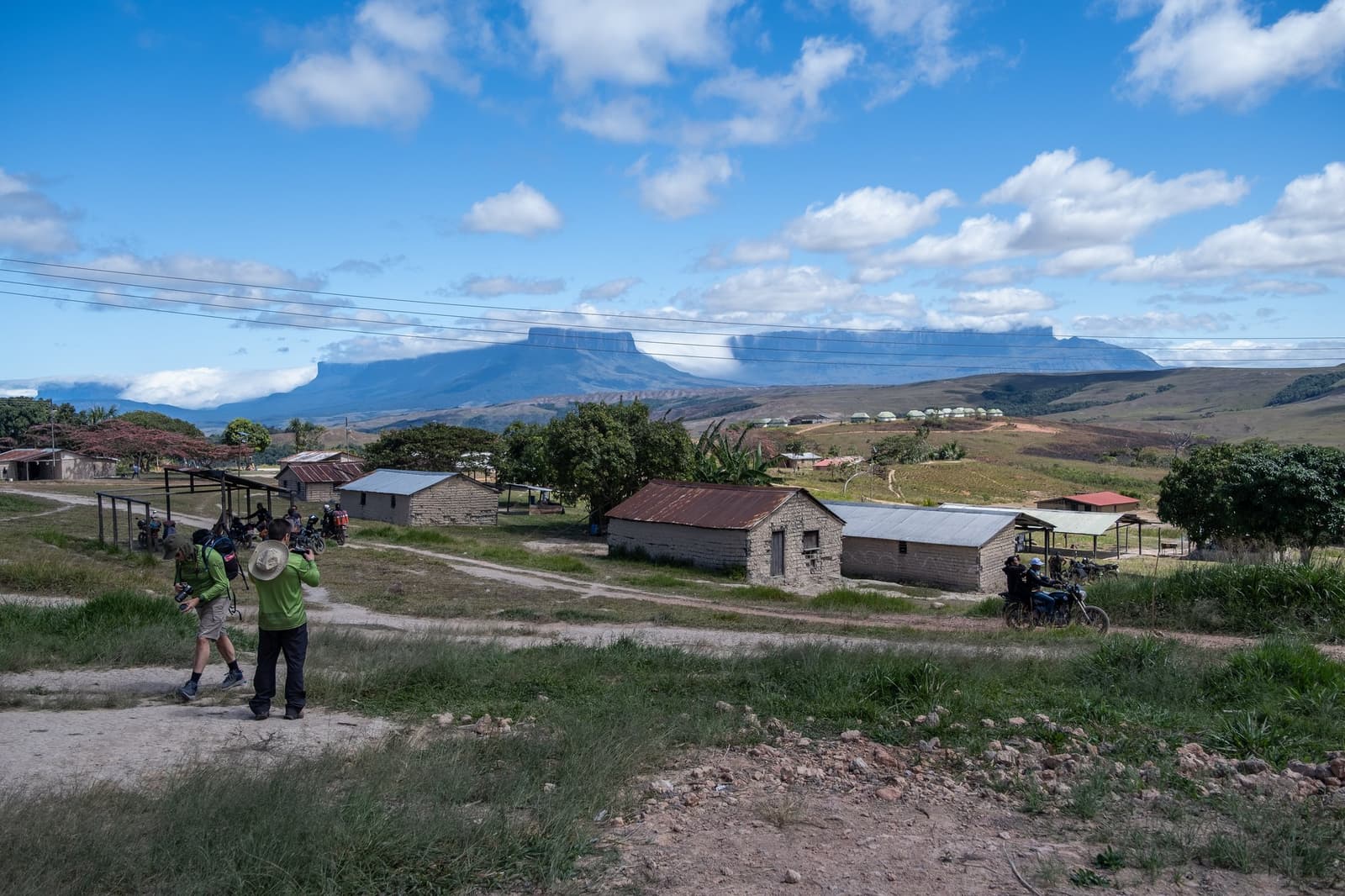 Paraitepuy: the last village before the mountain takes over. Two photographers with cameras photograph the Paraitepuy village on a mount roraima expedition, with stone-and-mortar houses, a dirt road, and a flat-topped tepui rising dramatically on the horizon under a blue sky with scattered clouds