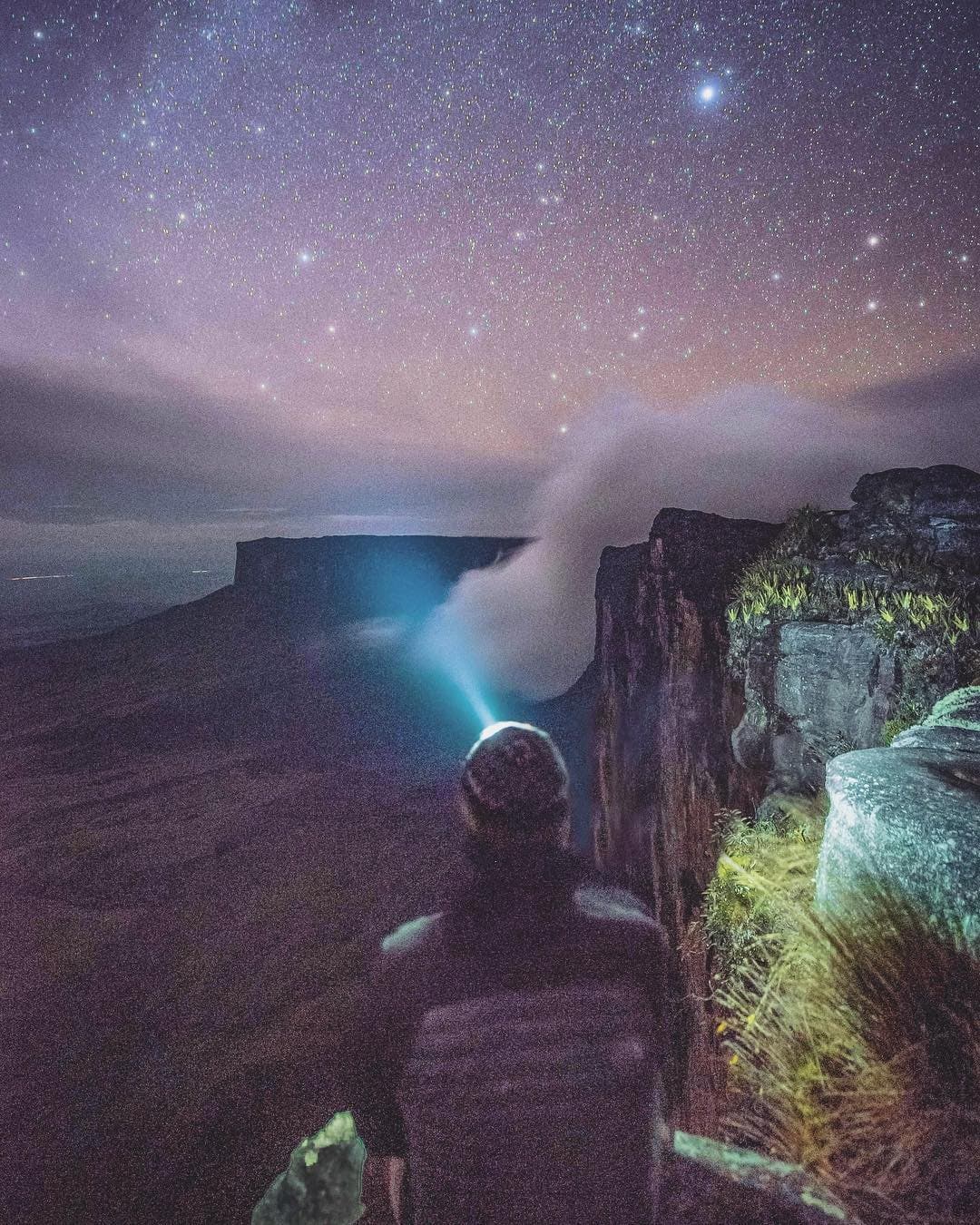 Some nights the mountain hands you the whole sky and asks nothing back. A hiker wearing a headlamp stands at the edge of a tepui cliff on a mount roraima expedition, gazing across a sea of mist toward a distant flat-topped mesa under a dense, star-filled sky