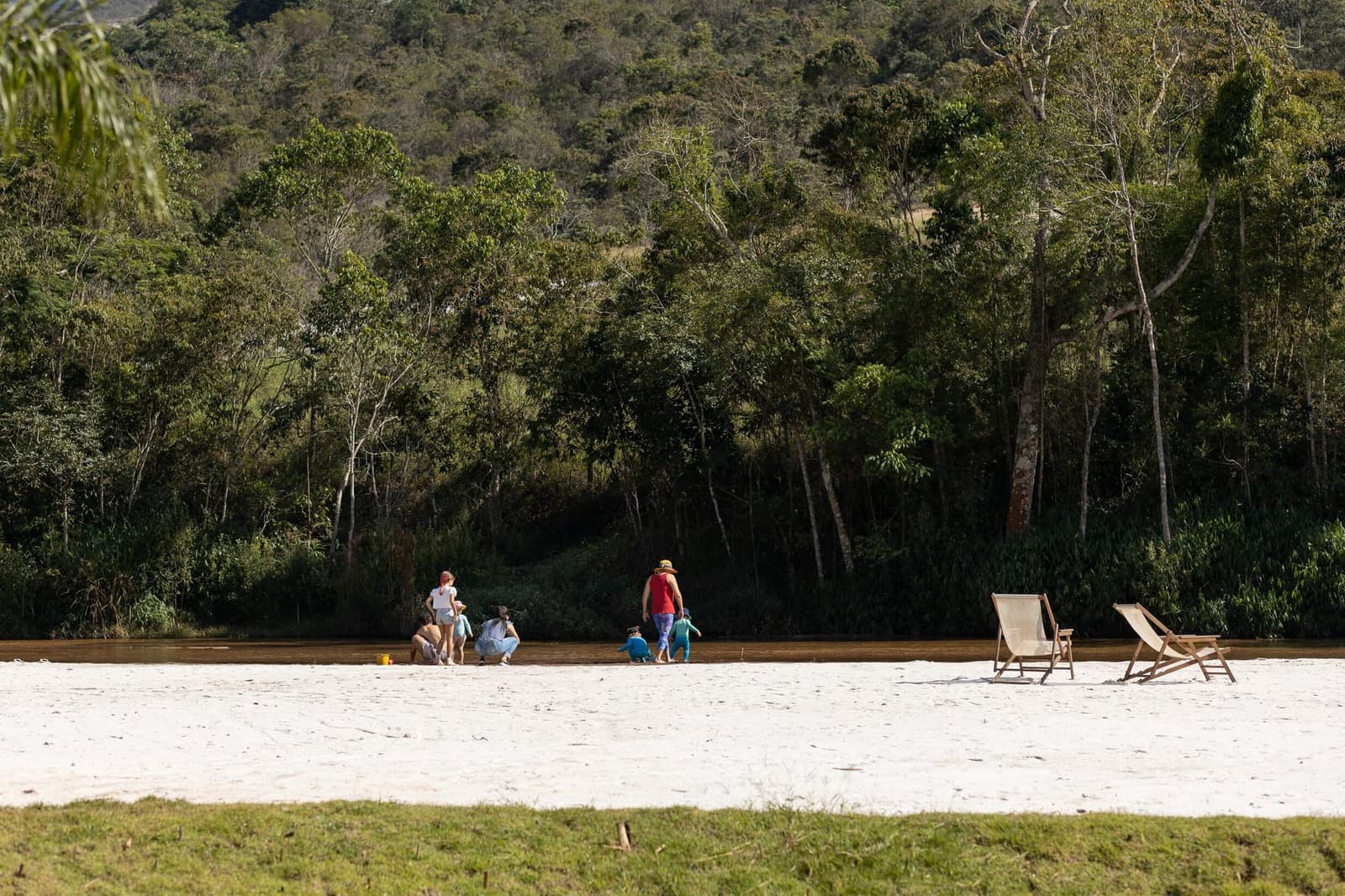 Sand, a slow river, and kids who found exactly what they needed. Families and children play at the edge of a sandy river beach at the Ibiti Project, with wooden sun loungers nearby and dense Atlantic Forest rising on the hillside behind