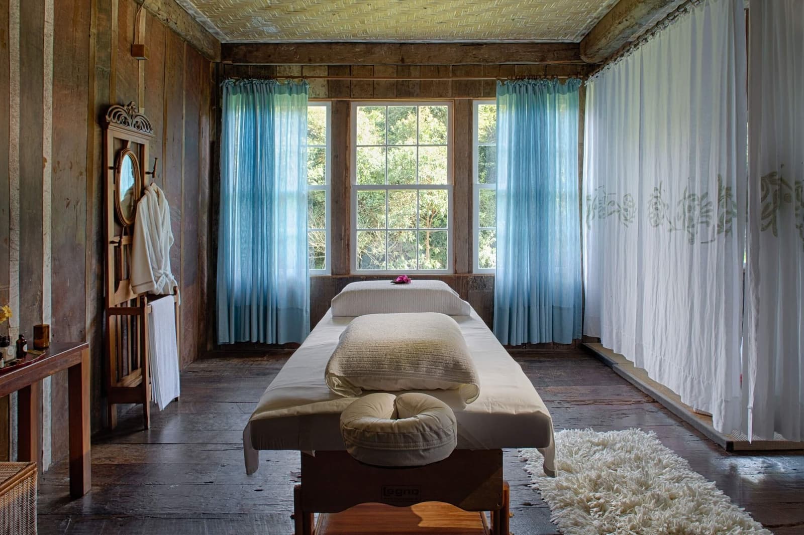 Quiet room, forest just outside the window, no agenda until tomorrow. A serene massage room at the Ibiti Project's Spa Raízes, with a padded treatment table, soft pillows, light blue sheer curtains, rustic wooden walls, and a white bathrobe hanging on a vintage stand beside a window overlooking green treetops