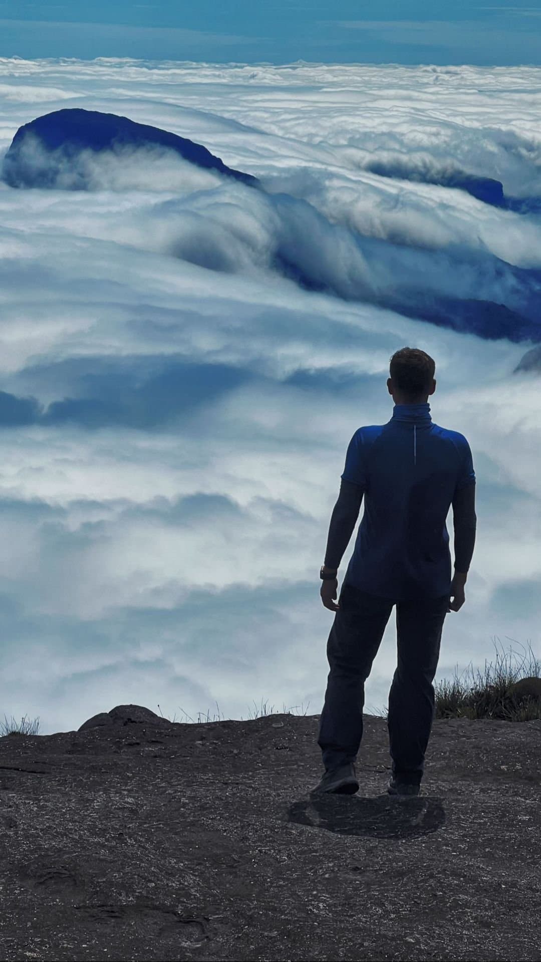 Standing above the clouds, and the whole world goes quiet. A hiker stands on a rocky summit ledge during the mount roraima expedition, looking out over a vast sea of clouds with dark tepui peaks barely breaking through the white below