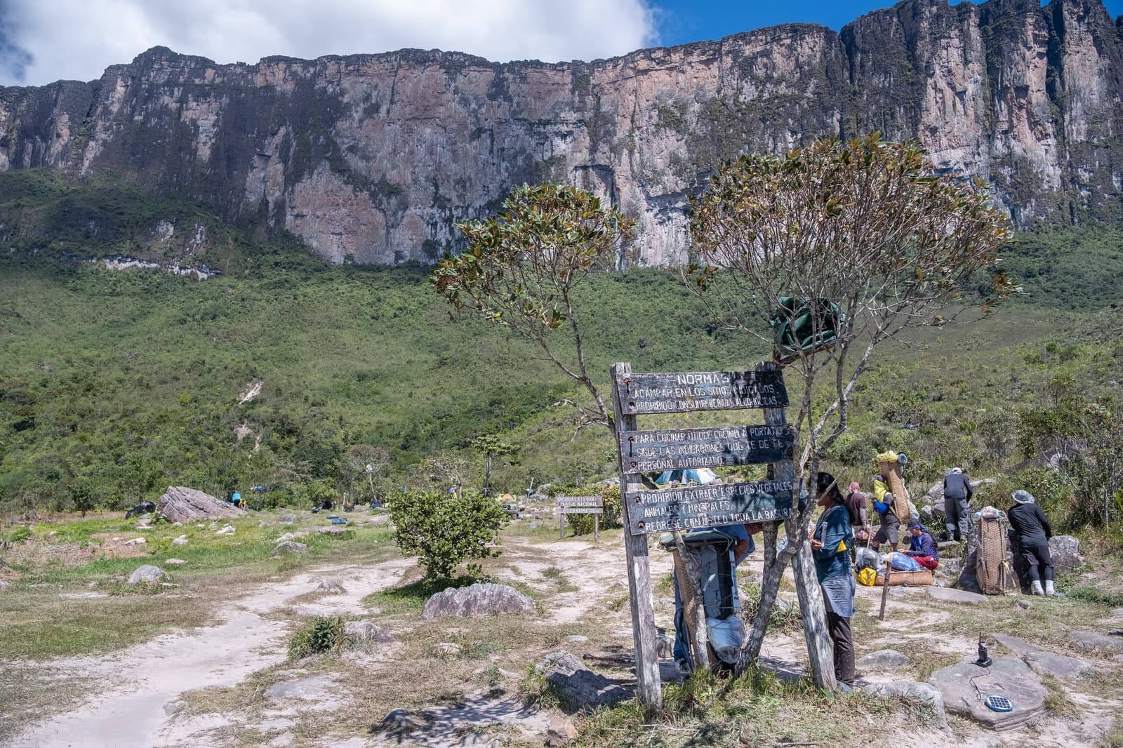 Rules posted, packs loaded, and the cliff already making its presence felt. A wooden rules sign reading 'Normas' stands at the base camp of a mount roraima expedition, with porters loading traditional backpack frames and the sheer tepui cliff wall rising behind the camp