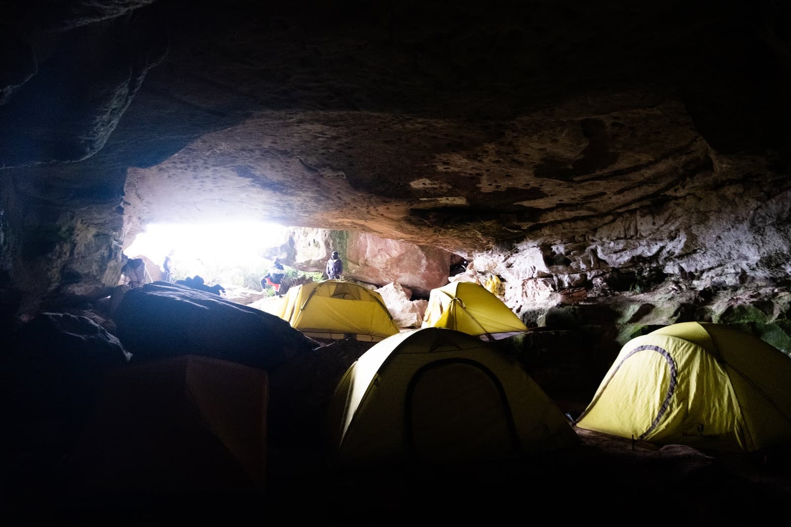 Stone roof overhead, no tent pegs required. Coati Camp earns its reputation. Four yellow dome tents pitched inside a large rock shelter cave during a mount roraima expedition, with daylight and silhouetted trekkers visible through the low cave opening