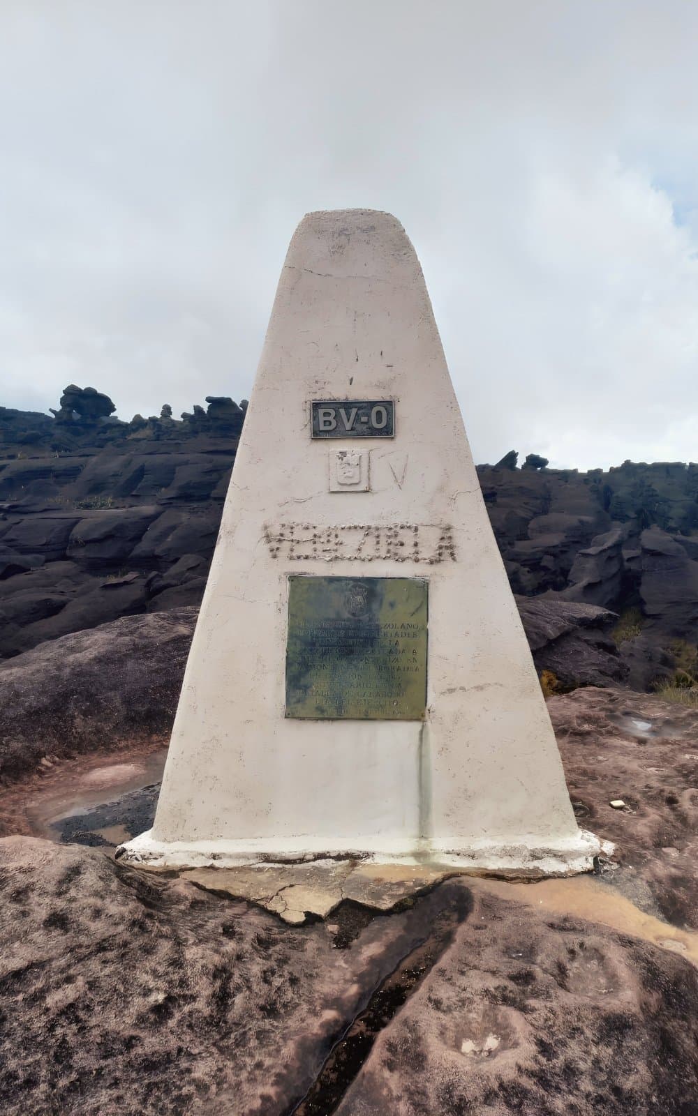 Three countries, one concrete obelisk, and a very long walk to get here. White pyramidal border marker labeled BV-0 and Venezuela standing on ancient dark rock at the Triple Point on the mount roraima expedition, where Venezuela, Brazil, and Guyana meet