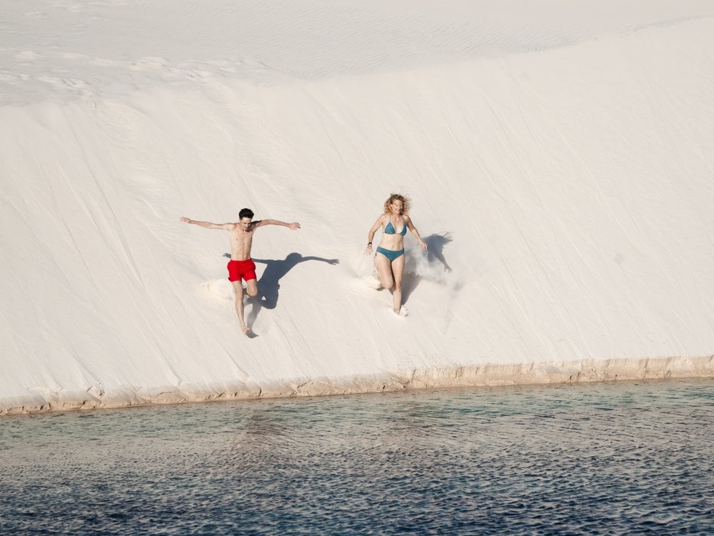 In search of an authentic experience far from mass tourism, French travelers Morgane and Guillaume opted for the classic cross-country hike. (Credit: Isadora Sá) Two travelers running down a white dune toward a lagoon in Lençóis Maranhenses