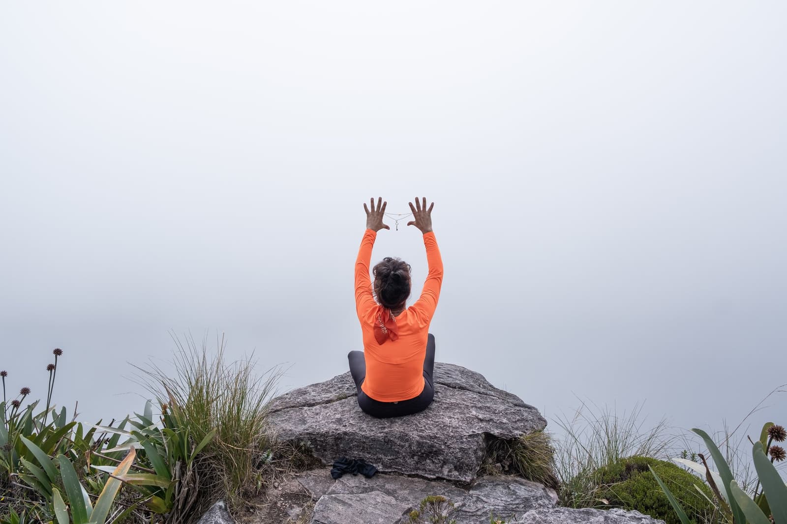 Fog as far as the eye can see, and honestly, that is enough. A person in an orange long-sleeve top sits cross-legged on a rock outcrop at the top of the mount roraima expedition route, arms raised above their head, surrounded by high-altitude vegetation and thick mist