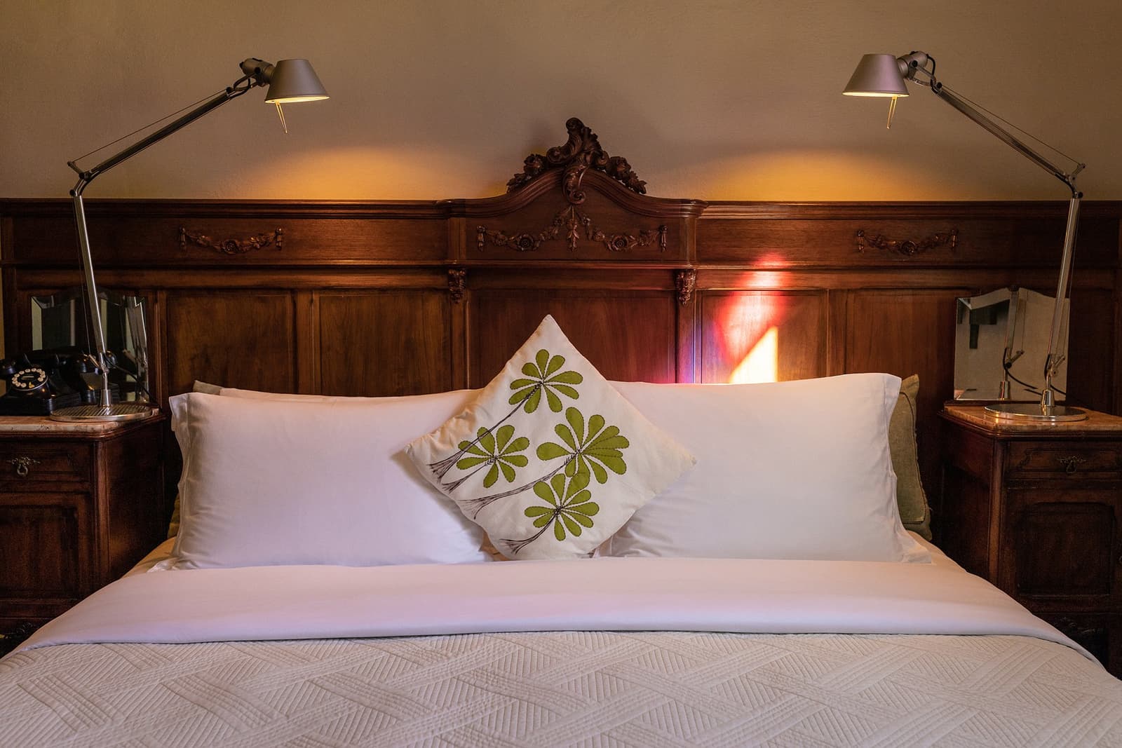 Antique headboard, fresh linen, the lodge earns its rest. Photo: Henrique Mangeon A king-size bed with crisp white linen and a leaf-patterned cushion sits beneath an ornately carved dark-wood headboard flanked by adjustable reading lamps at the Ibiti Project's Engenho Lodge