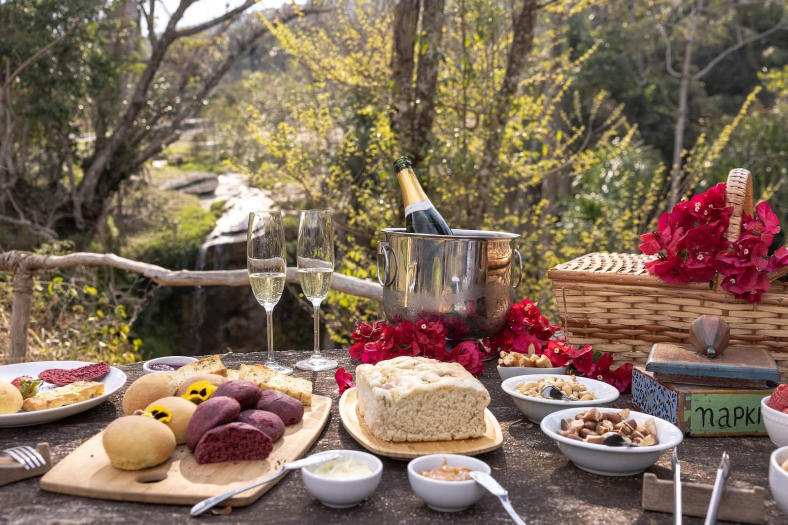 Champagne in the forest. Someone here knows how to do lunch right. An outdoor picnic table at the Ibiti Project laid with artisan breads, mixed nuts, dipping sauces, two champagne flutes, and a bottle chilling in an ice bucket, decorated with red bougainvillea flowers and a wicker basket, with leafy trees and a small waterfall visible in the background