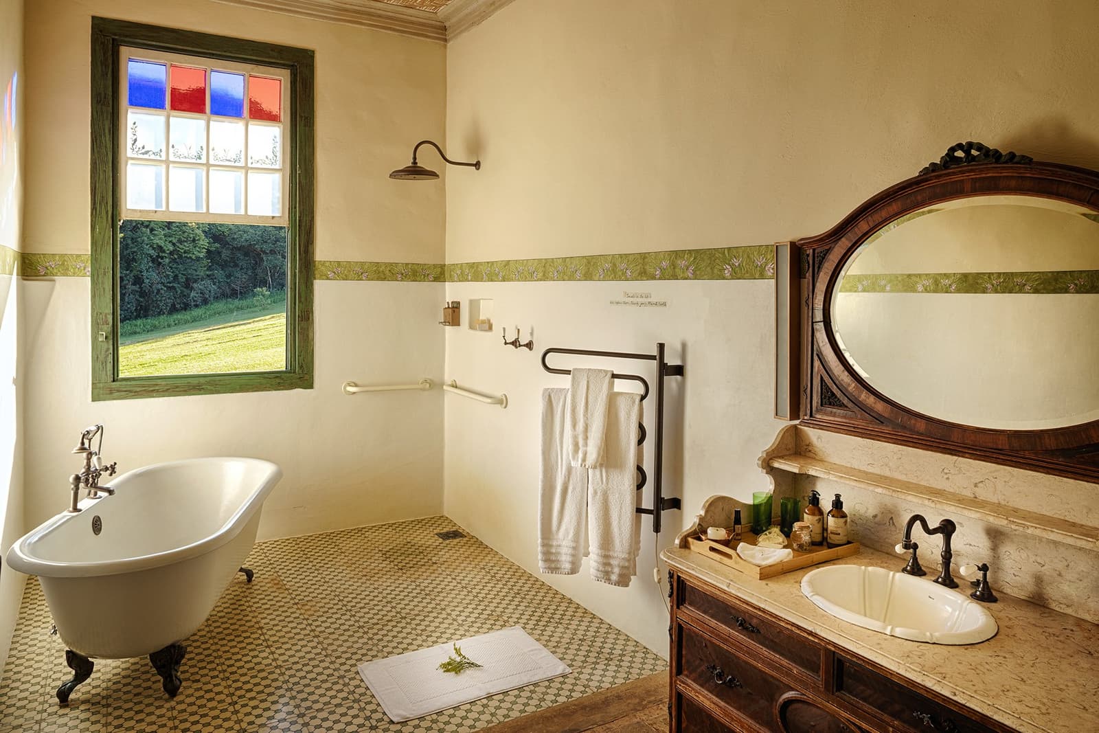 A clawfoot tub, a garden outside, and nowhere you need to be. Photo: Henrique Mangeon Clawfoot bathtub beside a stained-glass window with garden views in an ibiti project lodge bathroom, featuring a carved wood vanity with marble counter and antique oval mirror