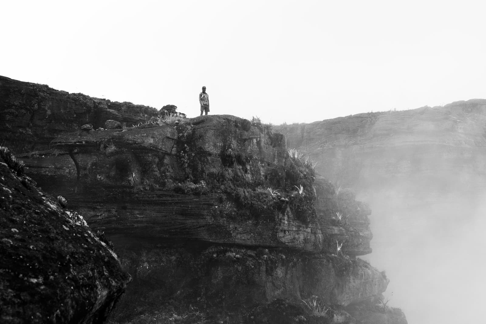 Just you, the cliff, and a cloud that hasn't decided where to go. A lone hiker stands at the edge of a moss-covered sandstone cliff on a mount roraima expedition, with dense mist rising from the canyon below and layered rock walls fading into white