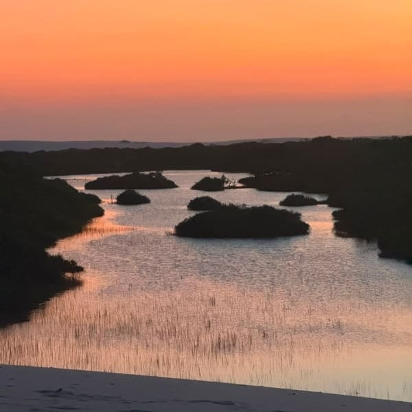 Orange sky reflected on a lagoon dotted with islands of vegetation, seen from a dune in Lençóis Maranhenses