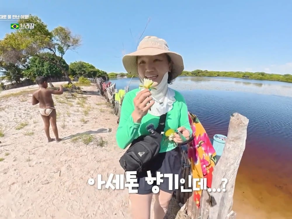 Photo: KBS Jihee Yeon holding small yellow flowers by the water, with a local child in the background