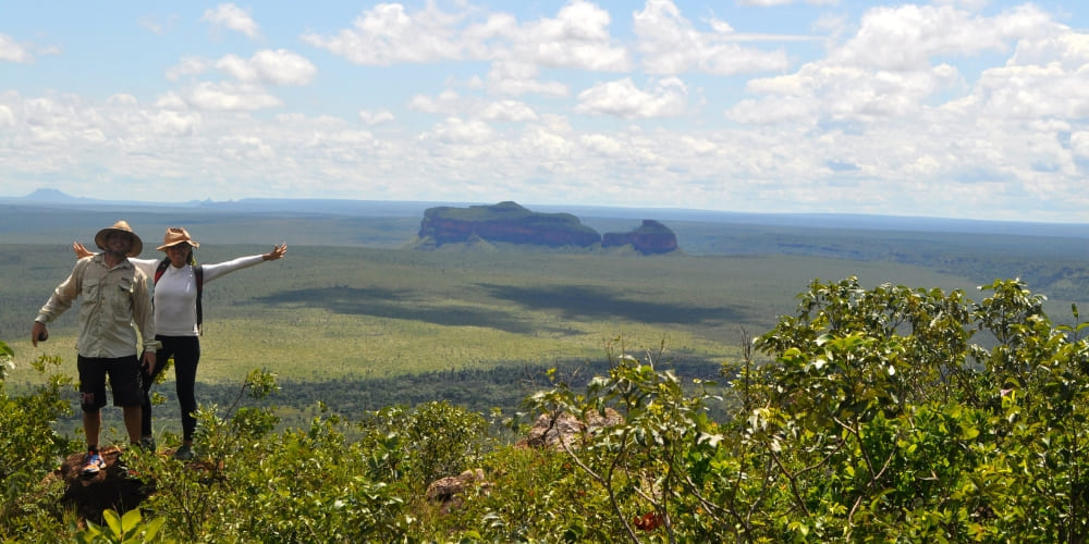 Two hikers stand on a lush green hilltop with their arms raised, enjoying a panoramic view of the Morro da Catedral rock formation.
