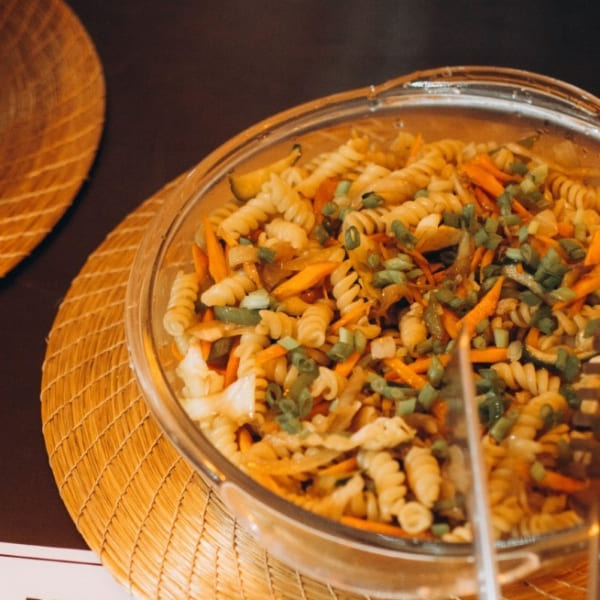 A glass serving bowl filled with rotini pasta salad, carrots, and green onions on a woven placemat, featured at a regional Jalapão restaurant buffet.
