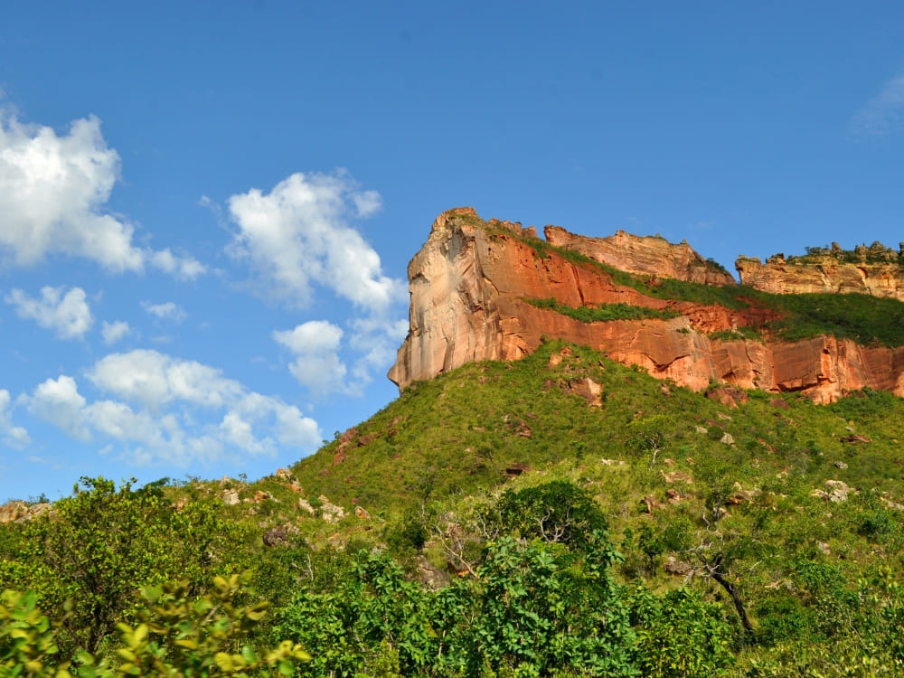The towering red sandstone rock face of Morro da Catedral rises above the green Cerrado landscape under a bright blue sky.