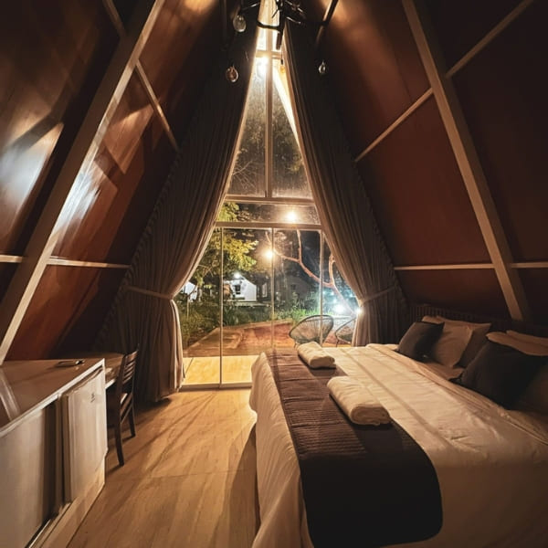 Inside a cozy A-frame chalet bedroom looking out through glass doors into the Jalapão forest at night.