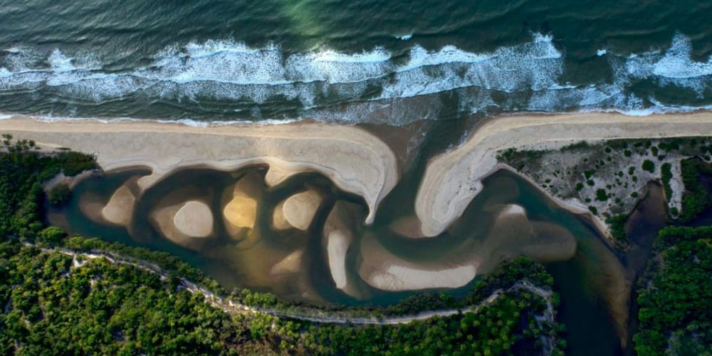 An aerial view shows a meandering river winding through sandbars and emptying into the ocean along the coast of Bahia.