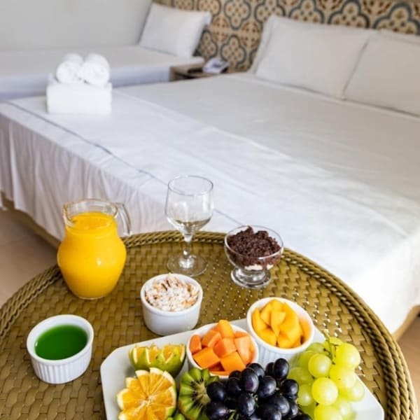A woven tray filled with fresh fruit, granola, juice, and coffee sits on a white hotel bed.
