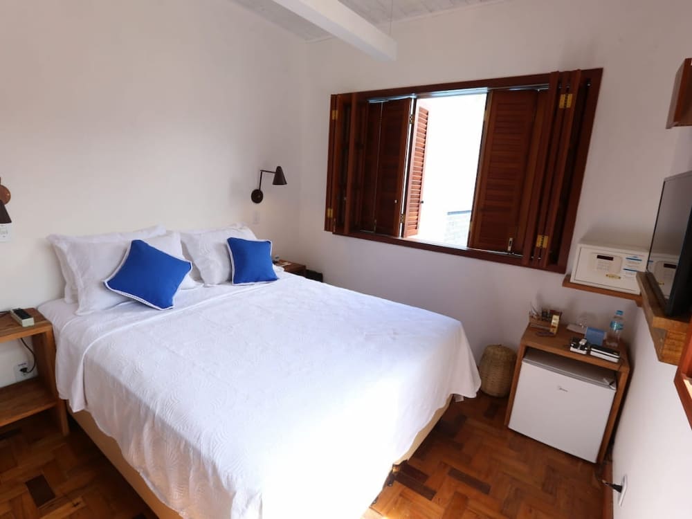 A cozy hotel room features a double bed with white linens and blue pillows, and a window with wooden shutters.