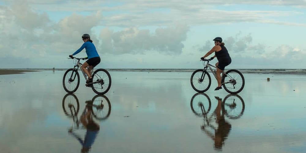 Two cyclists ride their bikes on a smooth, wet beach, creating perfect reflections of themselves on the sand.