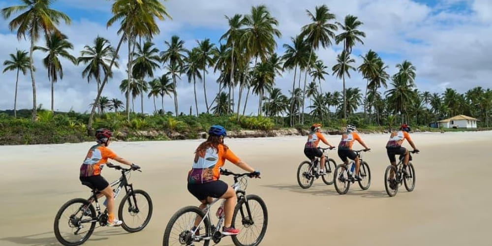 Four cyclists wearing orange shirts ride mountain bikes across a sun-kissed beach during a Bahia cycling tour.