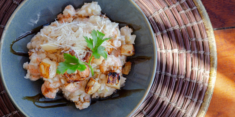 A close-up of a gourmet shrimp risotto dish topped with grated cheese and herbs, served in a grey bowl.