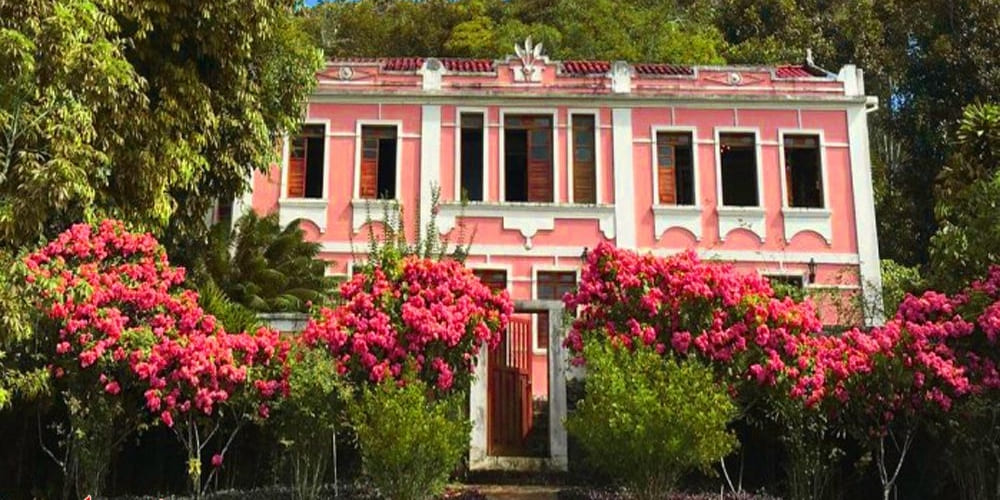 A bright pink colonial-style mansion with white trim is surrounded by lush green trees and vibrant pink blooming bushes at Fazenda Vila Rosa in Bahia.