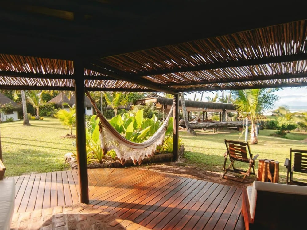 A white hammock hangs on a shaded wooden deck with lounge chairs, offering a relaxing view of a grassy yard and palm trees.