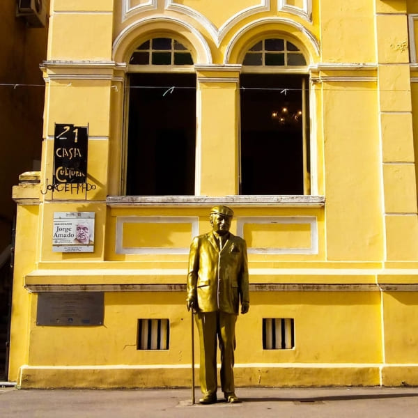 A golden statue stands in front of a bright yellow historic building in the city center of Ilhéus to highlight its connection with writer Jorge Amado.